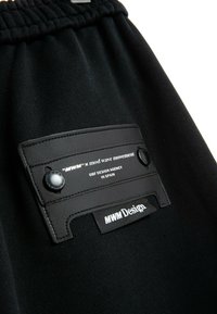 Pantalones cortos de tela negra con cintura elástica, una etiqueta de cuero texturizado con dos botones y detalles de marca impresos.