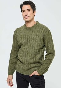 Pull en maille torsadée vert foncé avec un col rond, motif texturé et manches longues, associé à un pantalon noir. Coupe simple et décontractée.