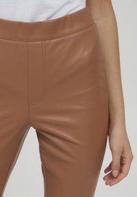 Bruine nep leren broek met een gladde textuur, hoge tailleband en nette stiksels. Het zijpaneel toont een getailleerde pasvorm en contouren.