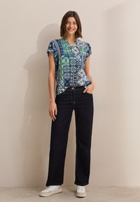 Top a maniche corte blu con motivo, decorato con accenti verdi e bianchi, abbinato a jeans a gamba larga blu scuro e sneakers bianche.