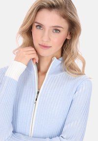 Jasnoniebieski sweter z zamkiem błyskawicznym, o pionowej, prążkowanej fakturze, z białymi akcentami na kołnierzu i mankietach oraz srebrnym zamkiem.