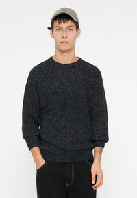 Joven vestido con una gorra gris, un suéter oscuro texturado y pantalones negros, con las manos en los bolsillos, de pie contra un fondo blanco liso.