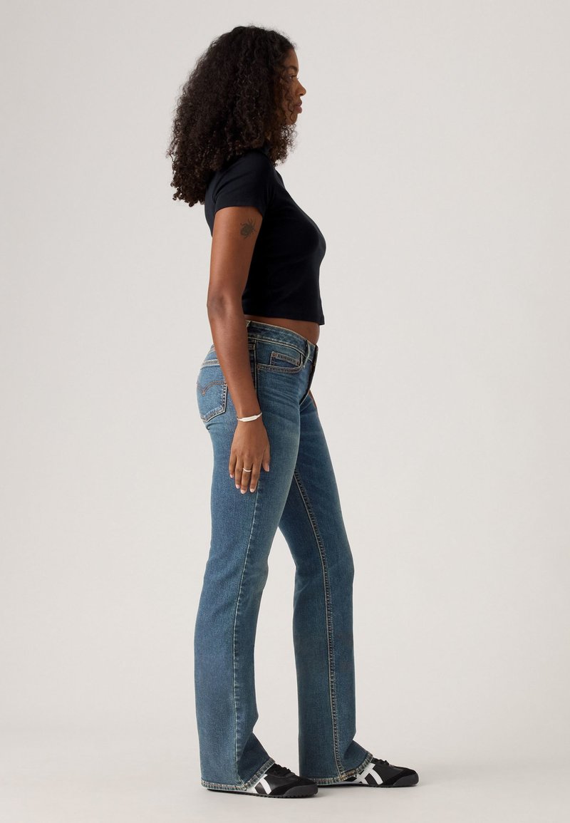 Black cropped t-shirt, light blue flared jeans, and black footwear with white accents. Curly hair, standing sideways against a neutral background.