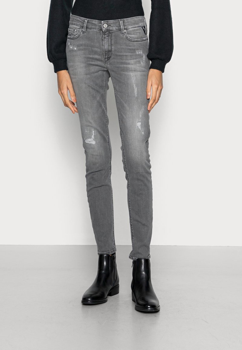 Slim-fit šedé džíny s prošlapanými detaily, designem s pěti kapsami a zapínáním na zip, doplněné černými kotníkovými botami. Hladká textura.