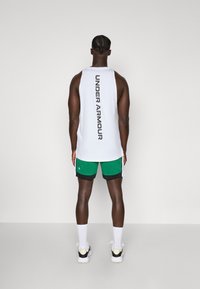 Camisola branca sem mangas com "UNDER ARMOUR" impressa verticalmente nas costas, combinada com calções de malha verdes e meias de desporto brancas.
