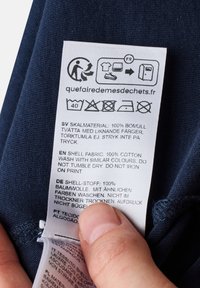 Bambukangas care label tummalla kankaalla; painettu teksti useilla kielillä antaa pesuohjeet ja materiaalitiedot. Käsi osittain näkyvissä.