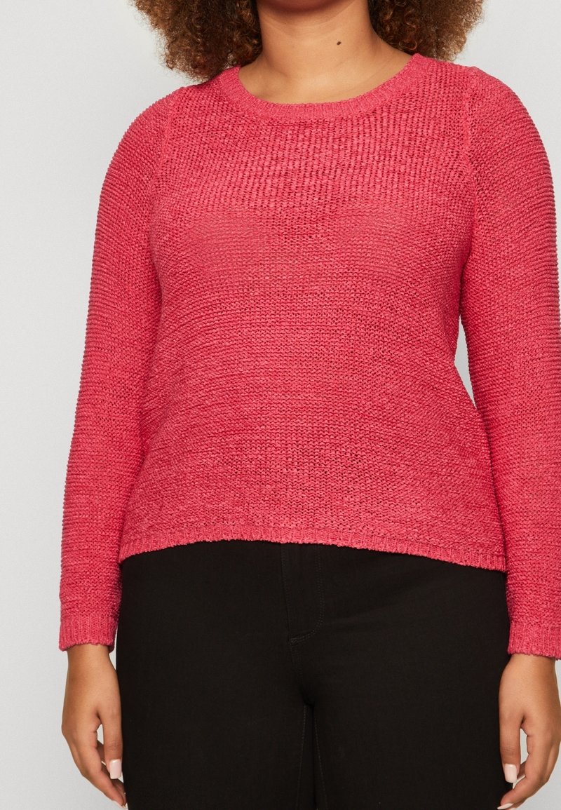 Femme portant un pull en tricot rose texturé à manches longues et un pantalon noir, debout avec les bras détendus le long du corps.