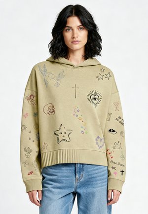 Donna che indossa una felpa con cappuccio beige con vari disegni ricamati tra cui stelle, uccelli, cuore con occhio, teschio e testo, abbinata a jeans blu.