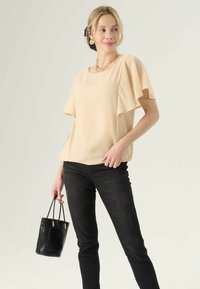 Perzikkleurige blouse met korte, transparante mouwen, gecombineerd met zwarte skinny jeans. Het model houdt een kleine textuur zwart handtas vast.