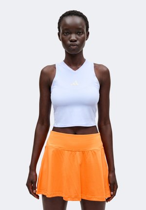 Γυναίκα που φοράει λευκό αμάνικο crop top και φωτεινή πορτοκαλί φούστα με καμπάνα, στέκεται μπροστά σε απλό φόντο.