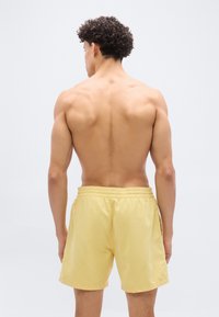 Νέος άνδρας με κατσαρά μαλλιά, γυμνός από τη μέση και πίσω, φοράει ανοιχτό κίτρινο σορτς πάνω σε λευκό φόντο.