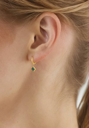 Pendiente de aro de oro con un pequeño colgante en forma de trébol de cuatro hojas verde, llevado en la oreja izquierda de una persona rubia.
