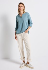 Vaaleansininen, pitkähihainen paita, jossa on V-pääntie ja rullatut hihat, yhdistettynä kermaisiin, löysiin housuihin. Huomiota herättävät taskut ja rento vauva.