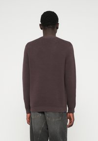 Άνδρας με κοντά σγουρά μαλλιά, φορώντας ένα ριγωτό μωβ πουλόβερ με μακριά μανίκια και σκούρα γκρι τζιν, γυρισμένος μακριά με φόντο απαλό φωτεινό χρώμα.