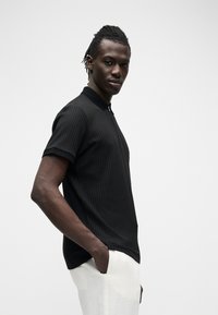 Homme aux dreadlocks portant un t-shirt noir côtelé à manches courtes et un pantalon blanc, debout avec une main dans la poche sur un fond uni.
