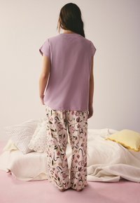 Personne debout, dos à la caméra, portant un haut lavande à manches courtes et un pantalon large à motifs floraux, près d'un lit avec des oreillers.