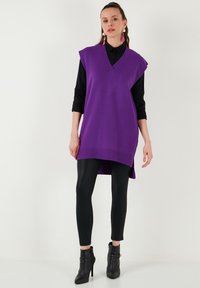 Lila kötött, ujjatlan tunika V-nyakkal, fekete leggings-szel és bokacsizmával párosítva. Bordás szegéllyel és oldalsó részekkel rendelkezik.