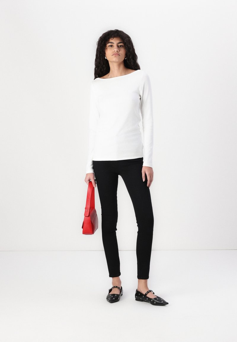 Langärmliges weißes Oberteil, schwarze enganliegende Jeans, schwarze flache Schuhe mit Ösen-Details, hält eine rote strukturierte Handtasche. Schlichte, klare Linien.