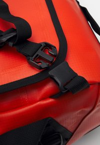 Röd vattentät väska med texturerad yta, svarta justerbara remmar och hållbara plastspännen. Har förstärkta sömmar som detaljer.