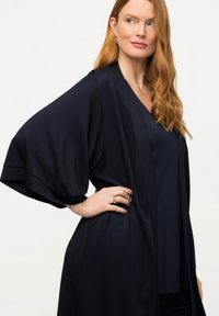 Tmavě modrý kimono stylový župan s širokými rukávy a volným designem. Vyrobený z lehkého materiálu s hladkou texturou. Obsahuje uvolněný střih.