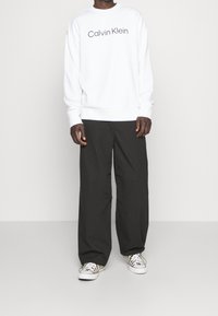 Witte crewneck sweatshirt met het "Calvin Klein" logo, gecombineerd met losse, donkere broeken en sneakers met een patroon. Casual, ontspannen pasvorm.