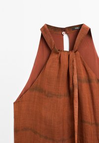 Rust-colored sleeveless top with a tie at the neck and a button closure at the back, featuring subtle fabric texture and pattern.