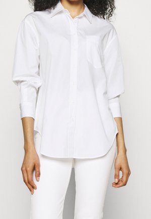 Personne portant une chemise blanche à manches longues boutonnée avec col et un pantalon blanc, debout devant un fond clair uni.