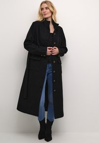Manteau long noir en mélange de laine, doté d'un col montant, d'une fermeture à boutons, de deux poches avant et d'une taille ceinturée. Le modèle porte un jean en denim et des bottes.