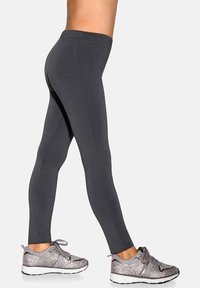 Osoba w ciemnoszarych dopasowanych legginsach i szarych błyszczących trampkach stoi z jedną nogą lekko wysuniętą do przodu na jednolitym tle.