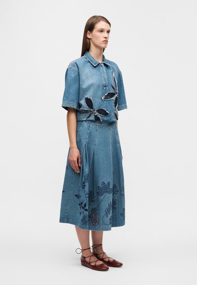 Ensemble haut et jupe en denim à manches courtes, avec broderies florales et plis. La tenue est bleue avec des accents foncés et des chaussures à lacets marron.