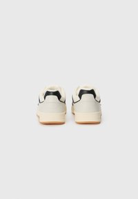 White and black sneakers with a smooth leather upper, padded collar, and a textured rubber sole in tan. Rear view, minimalist design.