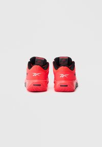 Ζευγάρι λαμπερών κόκκινων αθλητικών παπουτσιών με μαύρο εσωτερικό επένδυση και λευκό λογότυπο, εμφανιζόμενα από πίσω σε απλό λευκό φόντο.