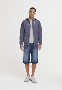 Sudadera gris con cremallera y cordones, llevada sobre una camiseta gris claro. Pantalones cortos de mezclilla, zapatillas grises. El modelo está de pie contra un fondo blanco liso.