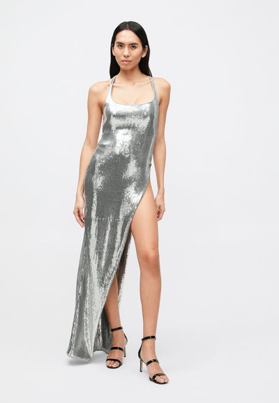 Strieborné trblietavé šaty s vysokým rozparkom a tenkými ramienkami, s priliehavým strihom. Doplnkové čierne sandále s remienkami.