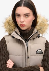 Beige Steppweste mit einer mit Fell gefütterten Kapuze. Verfügt über einen schwarzen Reißverschluss, ein Abzeichen auf der Brust und einen strukturierten braunen Strickpullover darunter.