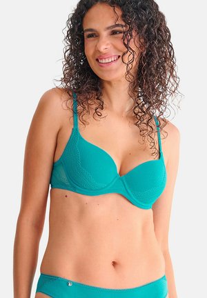 Woman with curly hair smiling, wearing a teal lace-accented bra and matching underwear against a plain light background.
