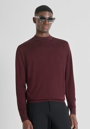 Hombre que lleva gafas de sol rectangulares, un suéter de manga larga burdeos y pantalones negros texturizados contra un fondo blanco.