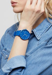 Orologio in silicone blu con quadrante circolare, numeri e un piccolo sotto-quadrante. Indossato su un polso con una camicia di jeans visibile sullo sfondo.