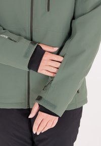 Grön softshell-jacka med en texturerad finish, med hög krage, dragkedjefickor och justerbara ärmslut med kardborre och elastik.