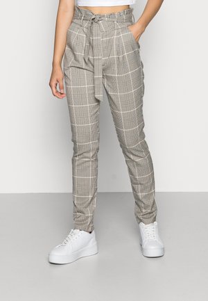 Vrouw draagt beige geruite broek met hoge taille en ceintuur gestrikt in de taille, gecombineerd met witte plateau sneakers, staand tegen een effen muur.