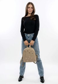 Zaino beige con accenti metallici circolari, materiale morbido, forma arrotondata, doppie cinghie per le spalle e tasca frontale con zip.