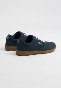 Zapatillas de lona azul marino con suela de goma, cordones blancos y detalle de logo. Textura suave con un diseño de caña baja y suela marrón en contraste.
