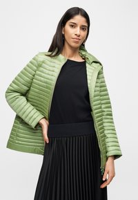 Γυναίκα που φοράει πράσινο ανοιχτό χρώμα μπουφάν με ραφές πάνω από μαύρη μπλούζα με μακριά μανίκια και μαύρη πλισέ φούστα, στέκεται με το ένα χέρι στη μέση της.