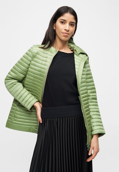Femme portant une veste matelassée vert clair par-dessus un haut noir à manches longues et une jupe plissée noire, posant avec une main sur la hanche.