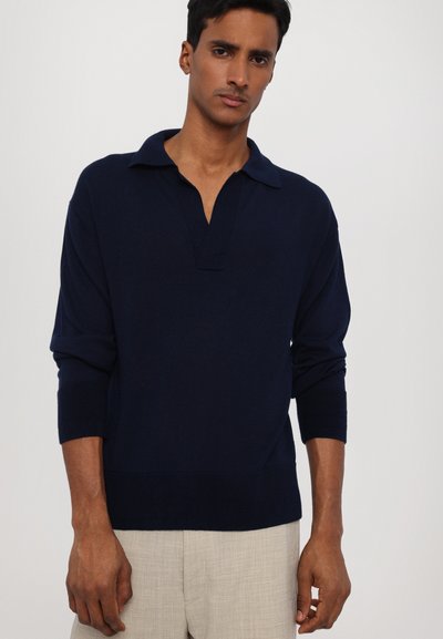 Pullover blu navy con scollo a collo e maniche corte. Texture liscia, polsini e orlo a costine. Indossato con pantaloni beige.