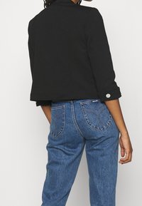 Persoon gekleed in een cropped zwart colbert met zilveren knoopdetails en high-waisted blauwe denim jeans met gebogen stiksels op de achterzakken.