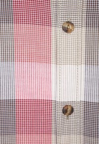 Geruite stof met beige, rode en bruine blokken. Opvallende details zijn een enkele bruine knop en fijne verticale lijnen in de weefsel.