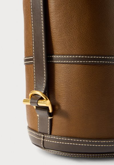 Saco de couro castanho com uma alça de couro mais escuro, detalhes em costura e um acabamento em metal dourado. Superfície texturizada e base arredondada.