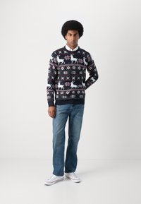 Sweater de malha em marinho com um padrão de rena e flocos de neve em branco, rosa e verde, combinado com jeans azul claro e ténis brancos.