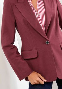 Bordeaux blazer gemaakt van textuurstof, met scherpe revers, een enkele zwarte knop en twee voorkruiszakken. Draag over een gestreept shirt.